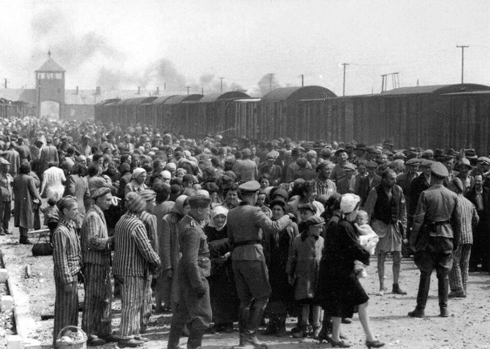 Auschwitz / autor: By Unknown photographer from the Auschwitz Erkennungsdienst. Several sources believe the photographer to have been SS officers Ernst Hoffmann or Bernhard Walter, who ran the Erkennungsdienst. - Yad Vashem: 'Jews undergoing selection on the ramp. Visible in the background is the famous entrance to the camp. Some veteran inmates are helping the new comers.', Domena publiczna, https://commons.wikimedia.org/w/index.php?curid=39226962