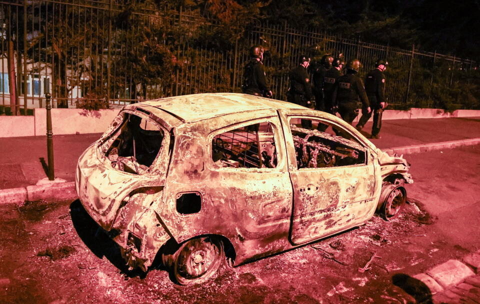 Policjanci idą obok spalonego samochodu zniszczonego podczas starć między protestującymi a policją w Nanterre, niedaleko Paryża / autor:  	PAP/EPA/MOHAMMED BADRA